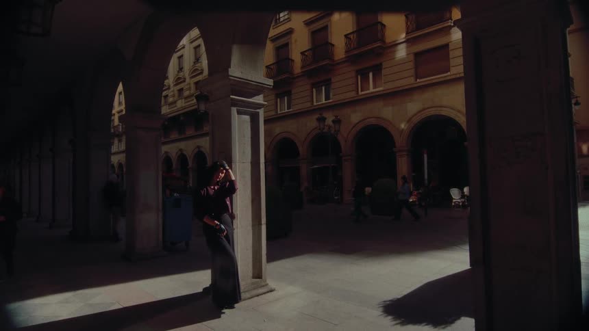 An elegant woman stands in the deep shadows of an arched colonnade, looking out into a sun-drenched European plaza