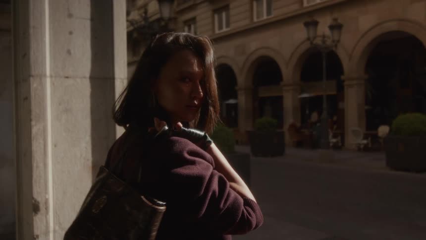 A mysterious woman in slow motion glances back over her shoulder while standing under a shadowy arcade on a sunny European street