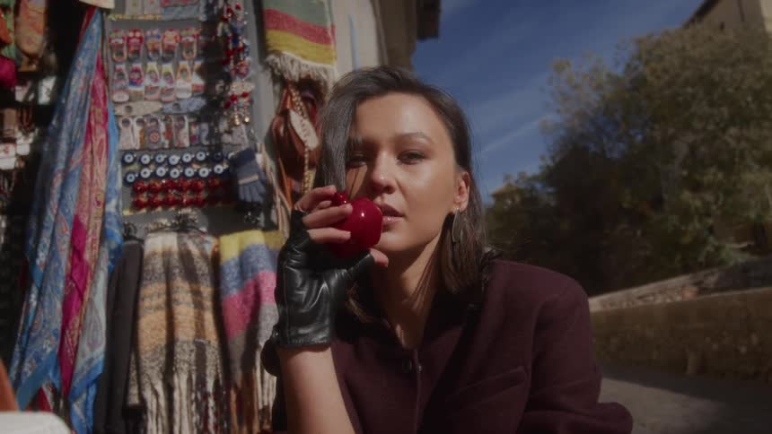 A stylish woman in a black leather glove holds a shiny red pomegranate and enjoys the sun at a colorful street market
