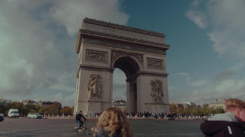 The historic Arc de Triomphe stands majestically in Paris as cars, cyclists, and pedestrians navigate the famous roundabout on a bright, cloudy day