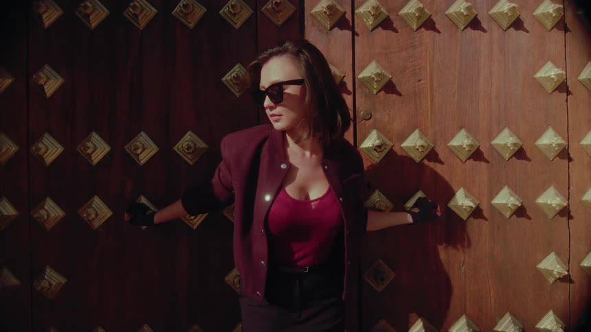 A fashionable woman in sunglasses stands against an old, studded wooden door as the wind blows her hair in slow motion