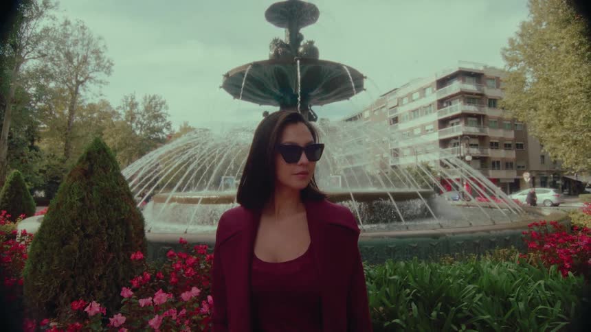 A stylish woman in sunglasses turns to look at the camera in slow motion while standing in front of a grand fountain in a city park