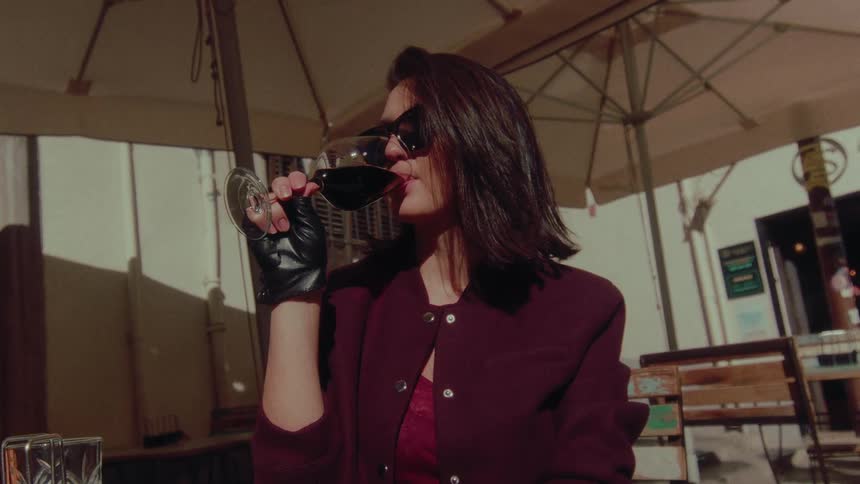 A stylish woman in sunglasses and leather gloves enjoys a glass of red wine at a sunny outdoor cafe in slow motion