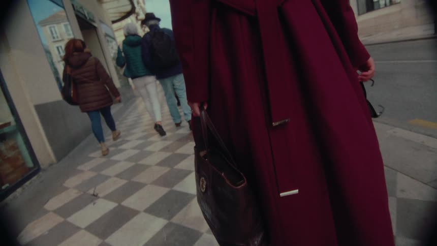 Vintage-style low-angle shot follows a woman in a long burgundy coat as she walks in slow motion along a checkered city sidewalk
