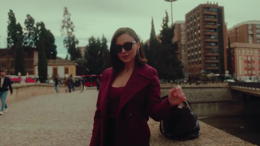 A fashionable young woman in a maroon coat puts on her sunglasses in slow motion on a cobblestone city walkway
