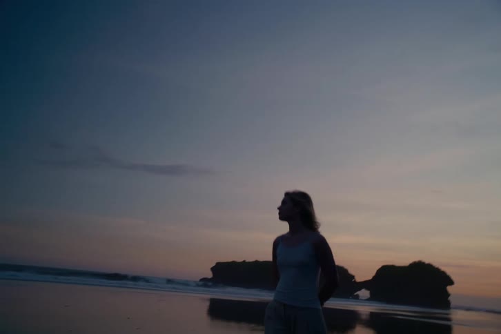 A woman in silhouette walks towards the camera on a reflective beach during a calm, beautiful sunset