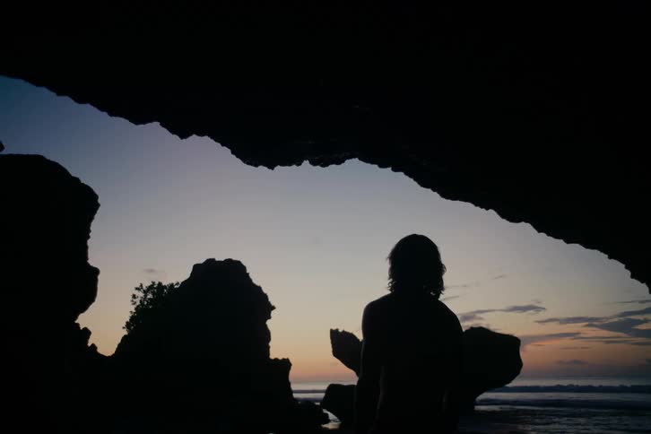 A silhouetted person watches the peaceful ocean and colorful sky at dusk from inside a dark coastal cave
