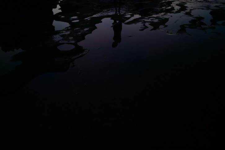A person's silhouetted reflection is distorted by ripples as they walk through dark water at twilight