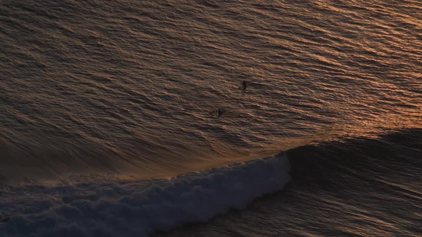 Slow-motion aerial view of a massive ocean wave building as two distant surfers wait patiently in the golden light of sunset