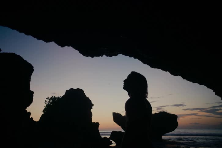A silhouetted person with long hair stands inside a coastal cave looking up at the serene twilight sky