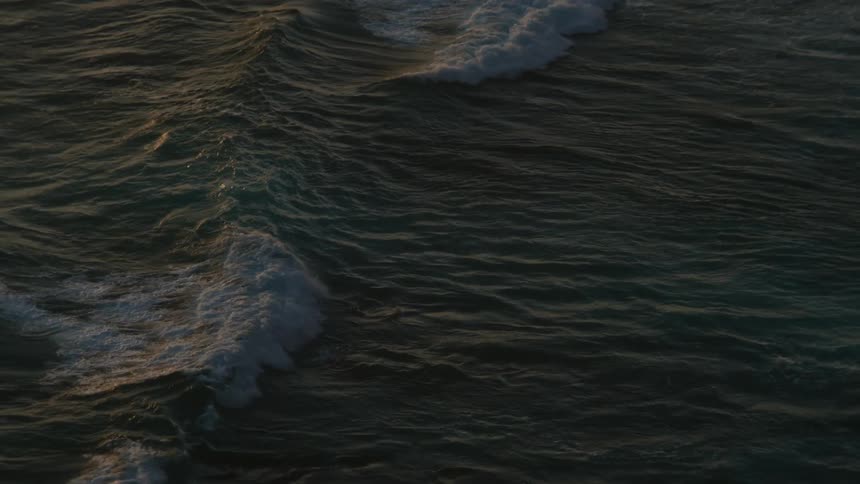 A top-down slow-motion view of dark, moody ocean waves rolling during sunset