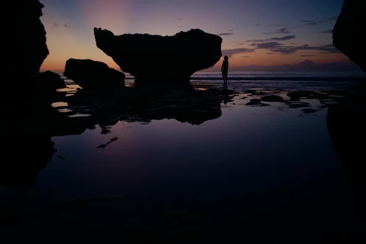 A solitary figure is silhouetted against a stunning purple and orange sunset sky on a rocky beach with still tide pools