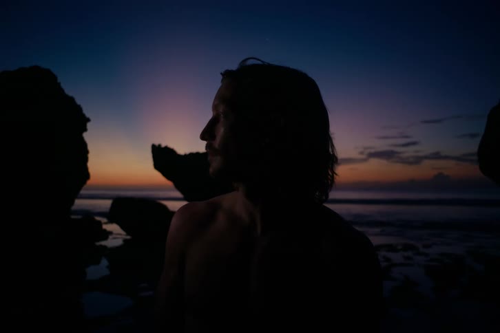 A shirtless man stands on a rocky beach as a flashing light reveals his profile against the twilight sky