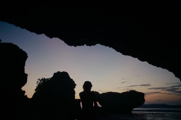 A silhouette of a person with long hair stands at the mouth of a seaside cave, peacefully watching the ocean waves during a serene twilight