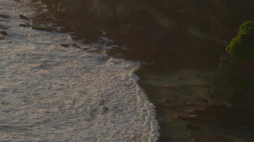 A serene, slow-motion high-angle view captures powerful ocean waves crashing against a rugged, rocky shoreline during the golden hour