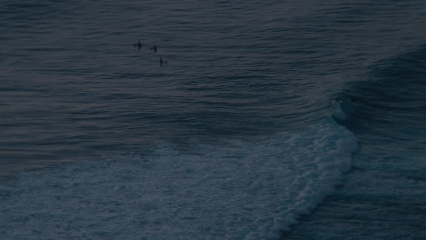 A distant, slow-motion shot captures a surfer riding a gentle wave as others wait in the twilight ocean