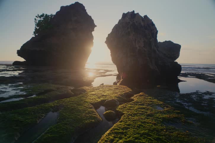 A serene sunrise peeks between two massive, silhouetted rock formations on a moss-covered shoreline at low tide