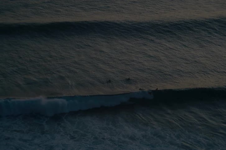 A serene, high-angle view of surfers waiting for the next big wave on the vast, dark ocean at dusk
