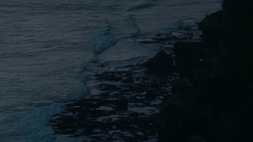 Slow motion waves crash against a rocky shoreline during a moody, dark evening