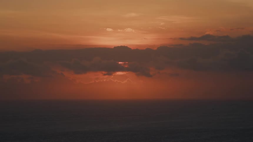A serene view of the sun setting over the ocean, with its vibrant orange glow peeking through dark, dramatic clouds