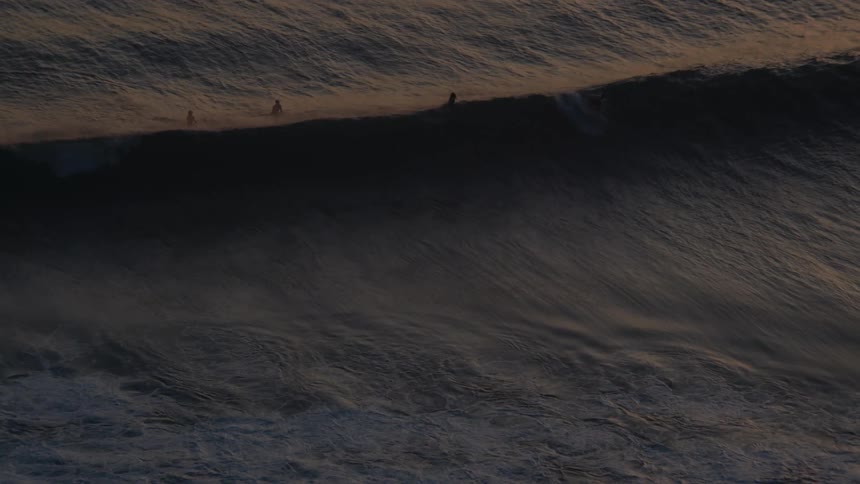 A serene aerial perspective captures surfers waiting patiently in the golden-hued ocean as large waves swell in slow motion