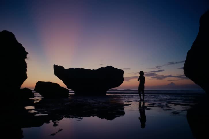 The silhouette of a person standing in a calm tide pool on a rocky beach is reflected in the water during a beautiful sunrise