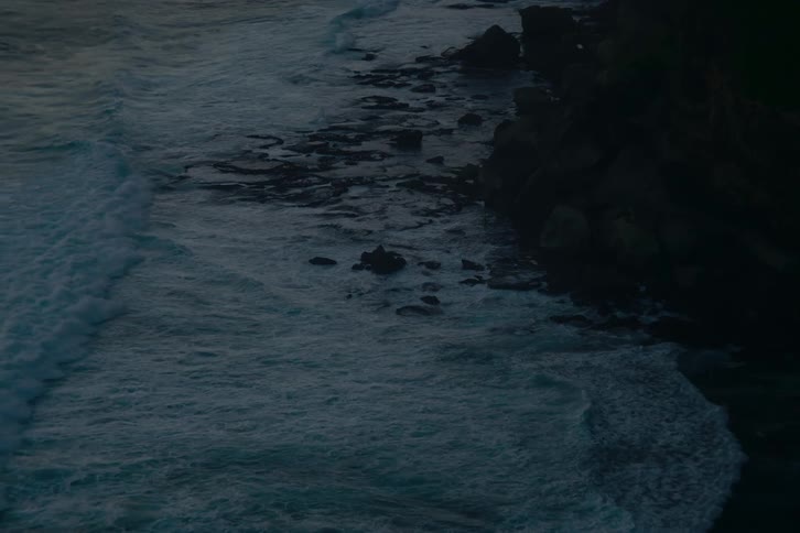 A moody, slow-motion, high-angle view captures powerful ocean waves crashing against a dark, rocky shoreline at dusk