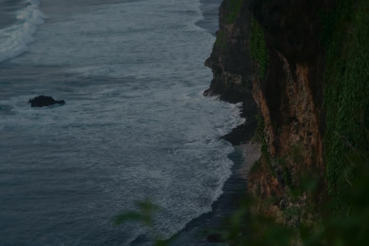 Moody, powerful ocean waves crash against the base of a sheer, verdant cliff