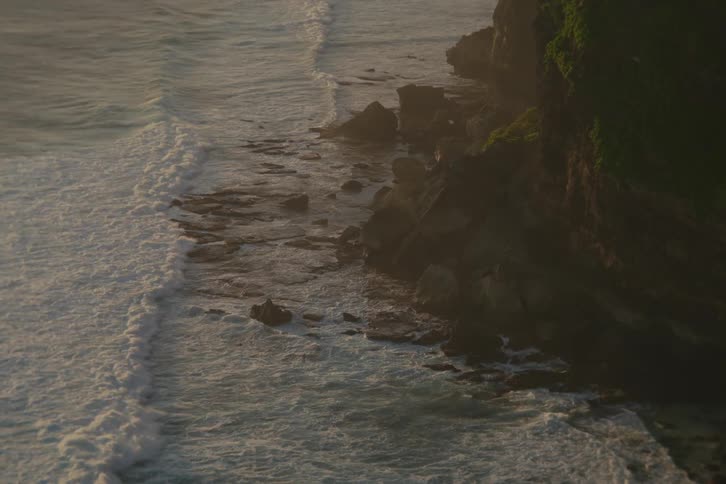 Gentle ocean waves crash against a rocky cliffside in slow motion during a hazy golden sunset