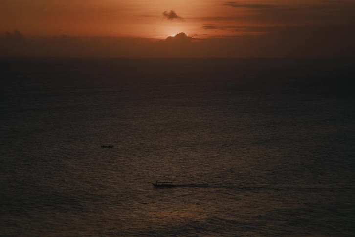 A serene, wide view of the vast ocean shows two small boats moving slowly under a beautiful orange sunset sky