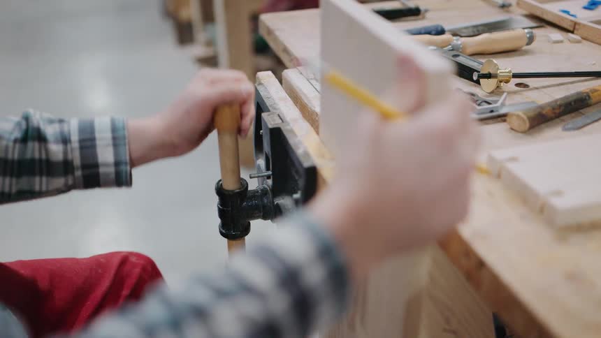 A woodworker tightens a vise on a wooden board at a workbench before marking it with a pencil