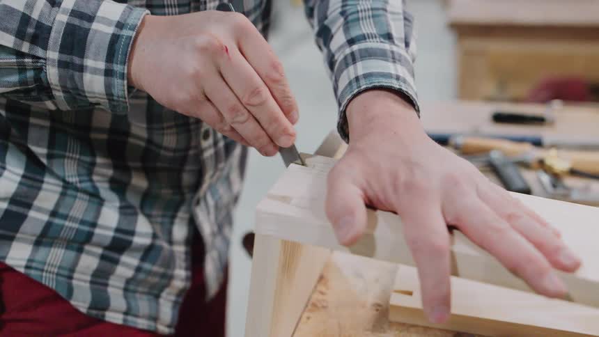A woodworker in a plaid shirt carefully uses a chisel to refine a dovetail joint on a piece of furniture in his workshop