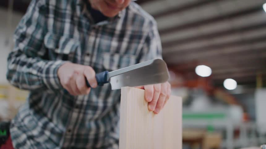 A focused carpenter in a plaid shirt and safety glasses carefully uses a Japanese pull saw to cut a piece of wood in a workshop