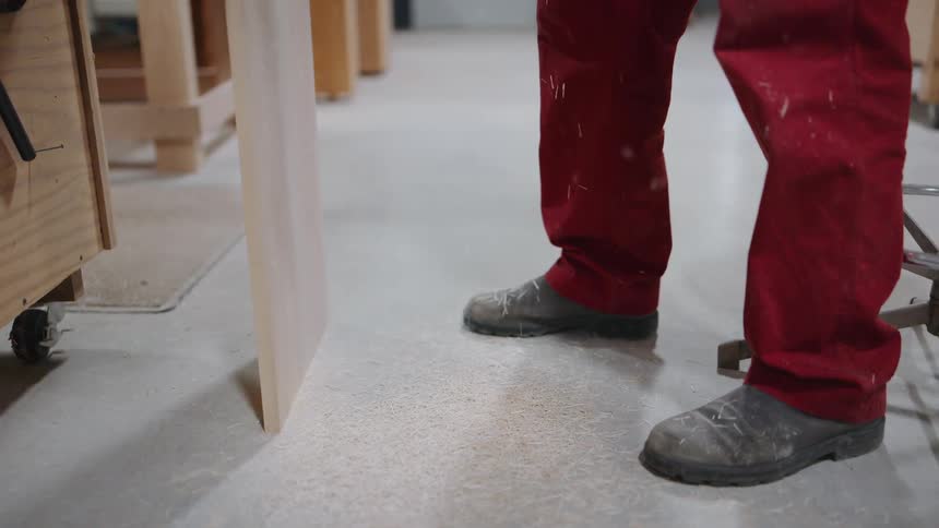 Sawdust falls in slow motion onto the floor and boots of a carpenter in a woodworking shop