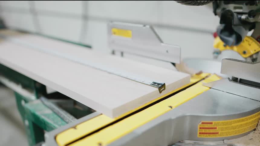 A woodworker uses a tape measure on a wooden plank resting on a miter saw in a workshop