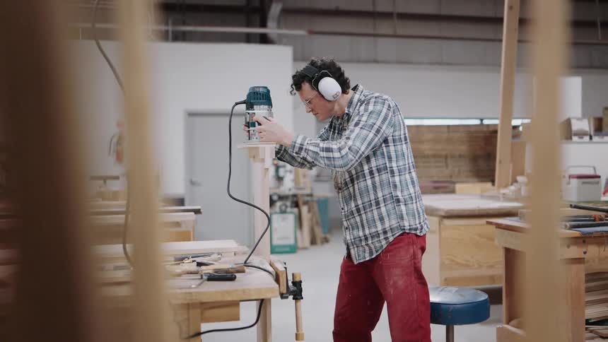 A professional woodworker in a workshop uses a power router to meticulously shape a piece of wood clamped to his bench
