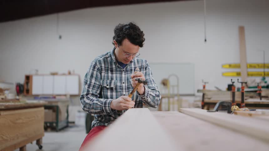 A skilled woodworker carefully uses a hammer and chisel to carve a long piece of wood in his workshop