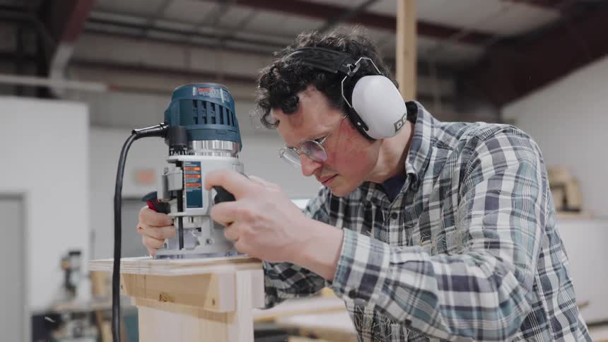 A professional woodworker wearing ear protection and glasses uses a plunge router to shape a piece of wood as sawdust flies in slow motion