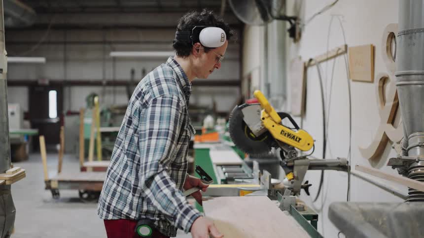 A focused woodworker in safety gear measures a wooden plank before making a cut with a miter saw in a workshop