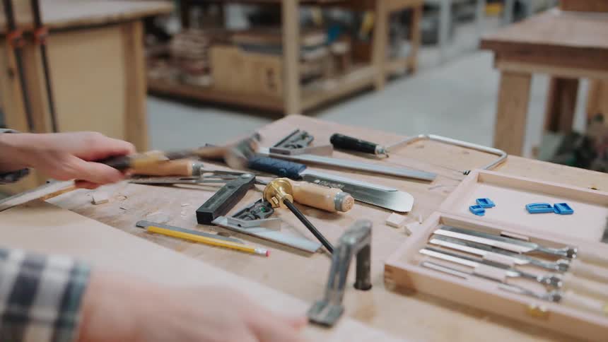 A carpenter in a workshop prepares to work by clamping a wooden plank to a workbench filled with various hand tools