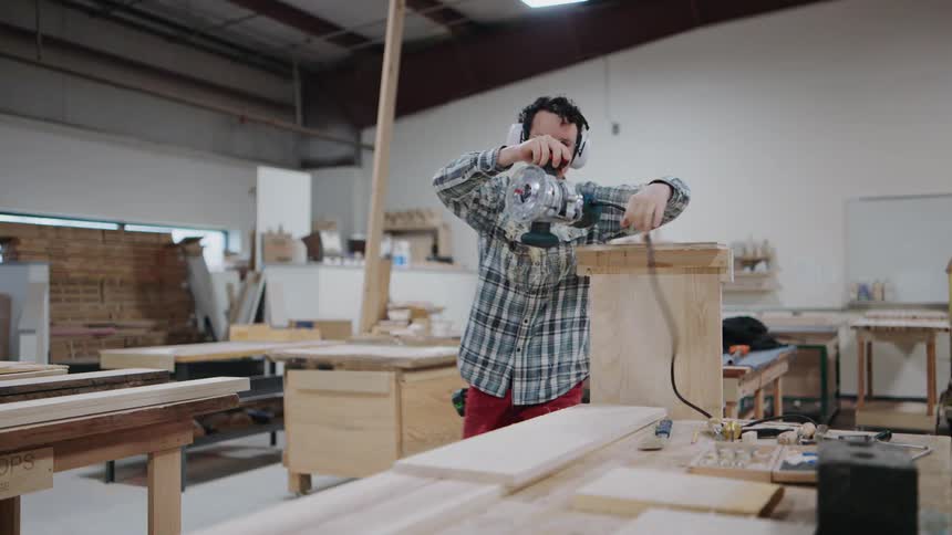 A skilled woodworker uses a router to craft furniture in his workshop before proudly smiling at the camera