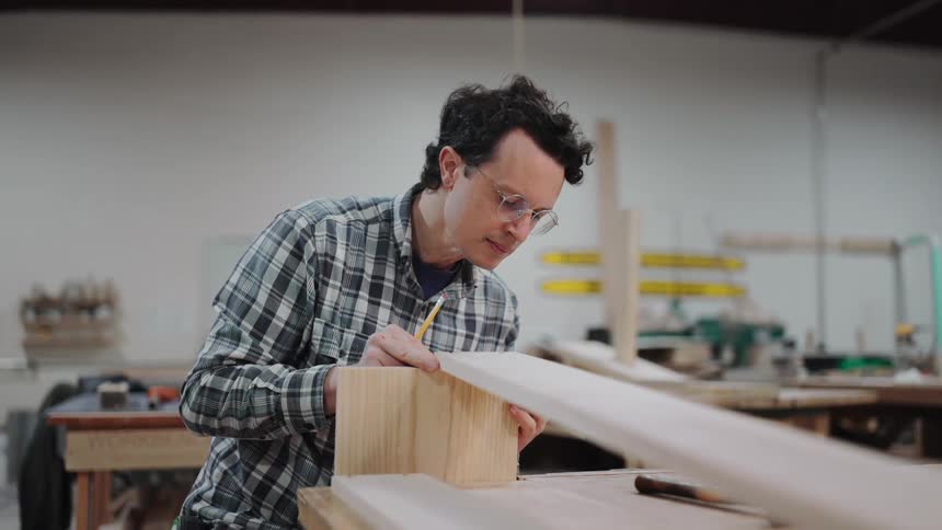 A focused craftsman in a plaid shirt meticulously measures and marks a piece of wood with a pencil in his workshop