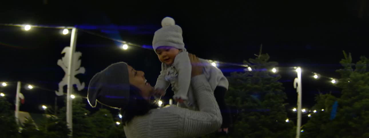 A woman holds up a baby while standing on a christmas tree lot at night