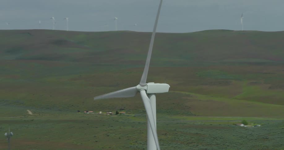 A dramatic aerial shot orbits a spinning wind turbine, revealing a sprawling wind farm generating clean energy across rolling green hills