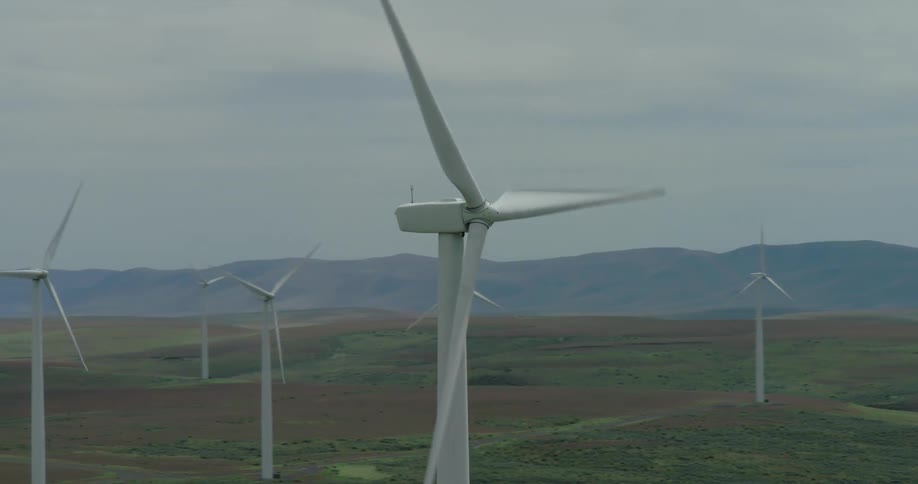 An aerial shot pulls back from a large spinning wind turbine to reveal a vast wind farm on a landscape of rolling hills under a cloudy sky