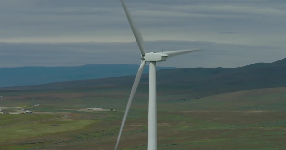 An aerial view pans around a large spinning wind turbine at a wind farm situated in a vast, hilly landscape on an overcast day