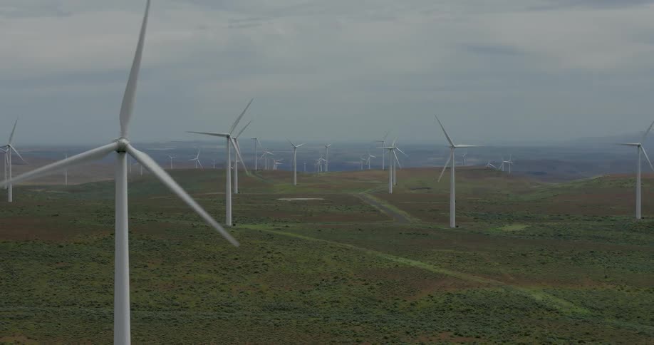 An aerial dolly shot passes by a spinning wind turbine, revealing a vast wind farm on a green, hilly landscape under an overcast sky
