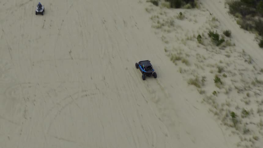 An aerial view captures a blue UTV kicking up sand as it drives up a massive dune
