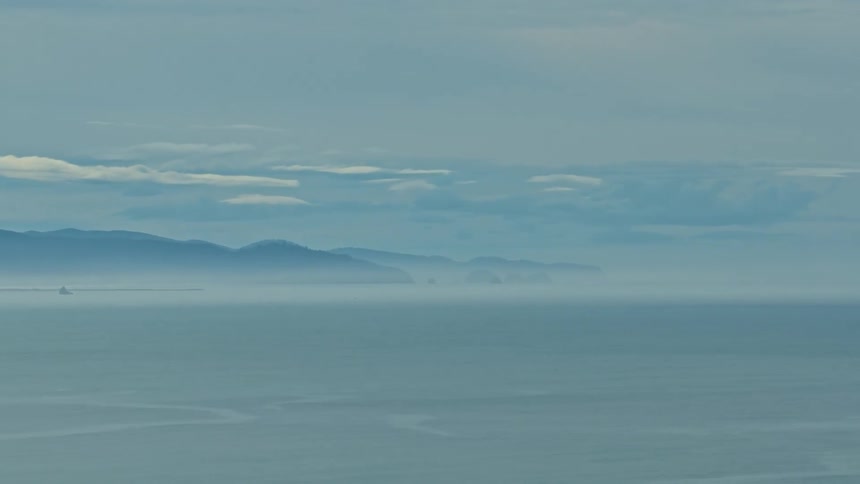 A calm, misty ocean stretches towards a hazy mountain coastline under an overcast sky