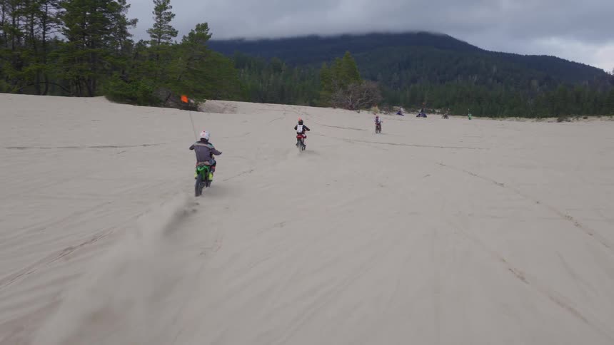 A group of friends enjoy an off-road adventure, riding dirt bikes across vast sand dunes with a forest and mountains in the background